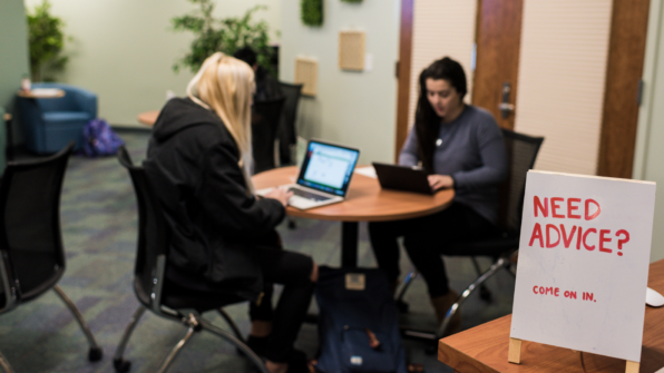 Students sit at a table with a sign in the foreground that reads "Need advice? Come on in!"