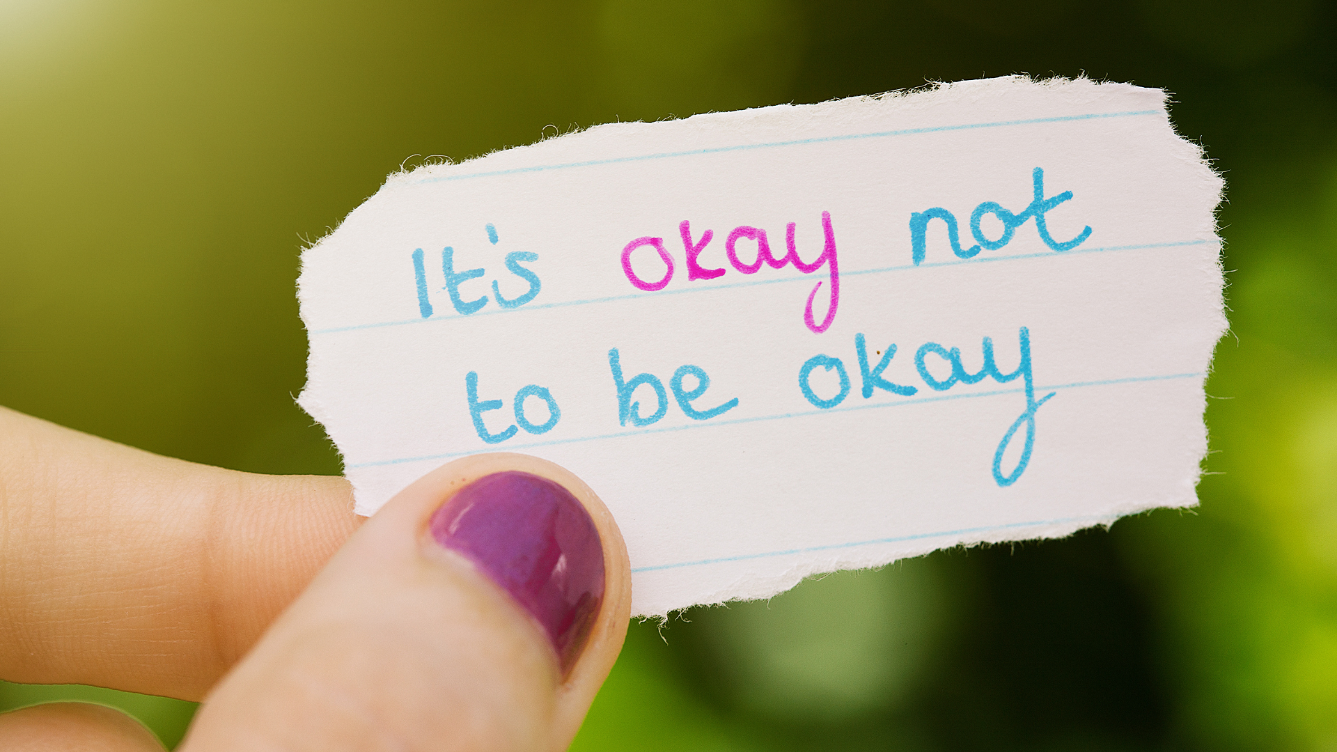 A piece of paper is held by a hand with purple nails that reads "It's okay no to be okay."