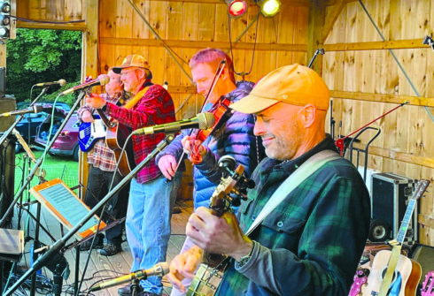 four people stand at microphones with various string instruments in their hands.