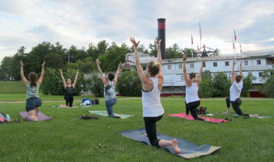 A group yoga class practices a pose outside on the lawn at Shelburne Museum, with the Ticonderoga ship in the background.