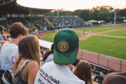 A student in a green and gold University of Vermont Catamounts hat watches a baseball game in the stands of Centennial Field as the sun sets behind the ballpark.