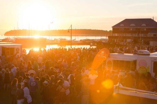 Hundreds of people gather at the food trucks and stages by the waterfront during sunset